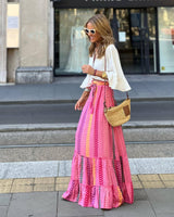 Donna con occhiali da sole indossa una gonna lunga a righe rosa e una blusa bianca, cammina in città con borsa di paglia. Moda estiva urbana.