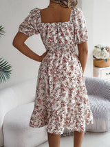 Donna in abito floreale estivo, maniche corte e scollo quadrato, in un salotto moderno con divano bianco e decorazioni naturali.
